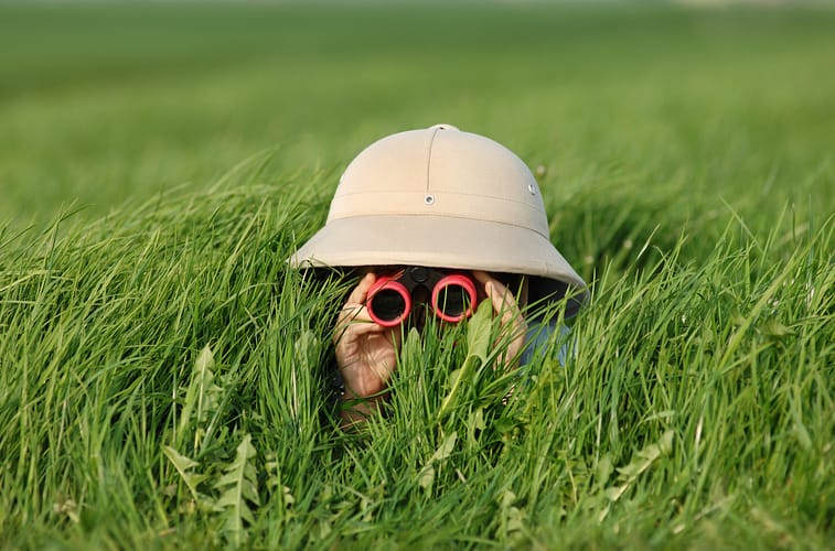 little boy in high grass searching for something using a pair of binoculars while he wears a safari helmet
