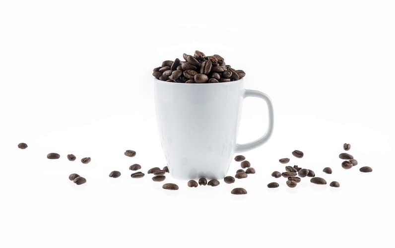 A white coffee mug on a white background that is overflowing with roasted coffee beans.
